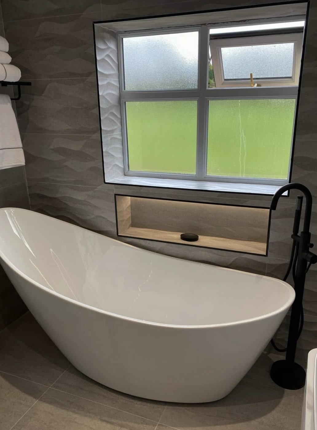 Slipper bath against a textured wave tile wall with a lit recess beneath the window